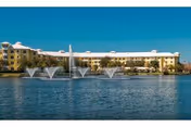 A large building complex named Cypress Cove situated behind a lake with multiple water fountains spraying water into the air under a clear blue sky.