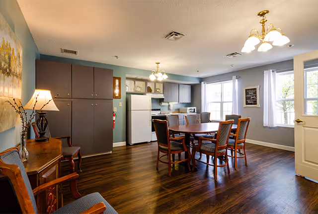 Bright common dining area with a round wooden table and chairs, a kitchenette with refrigerator and cabinets, and windows letting in natural light.