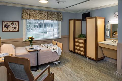 Private senior living bedroom with an adjustable hospital-style bed, chair, dresser and sink beneath a large window.