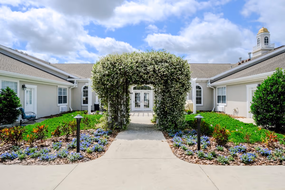 Outdoor view of a senior living facility entrance with a pathway leading through a lush green arch covered in white flowers. The pathway is flanked by flower beds with purple flowers and small shrubs. The building has light-colored walls, white-framed windows, and a cupola with a golden dome on the roof under a partly cloudy sky.