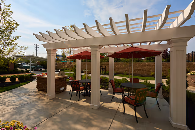 Outdoor patio area with a white pergola providing partial shade over several round tables with red umbrellas and cushioned chairs. There is a built-in grill station on the left side and landscaping with bushes and trees surrounding the paved area under a clear blue sky.