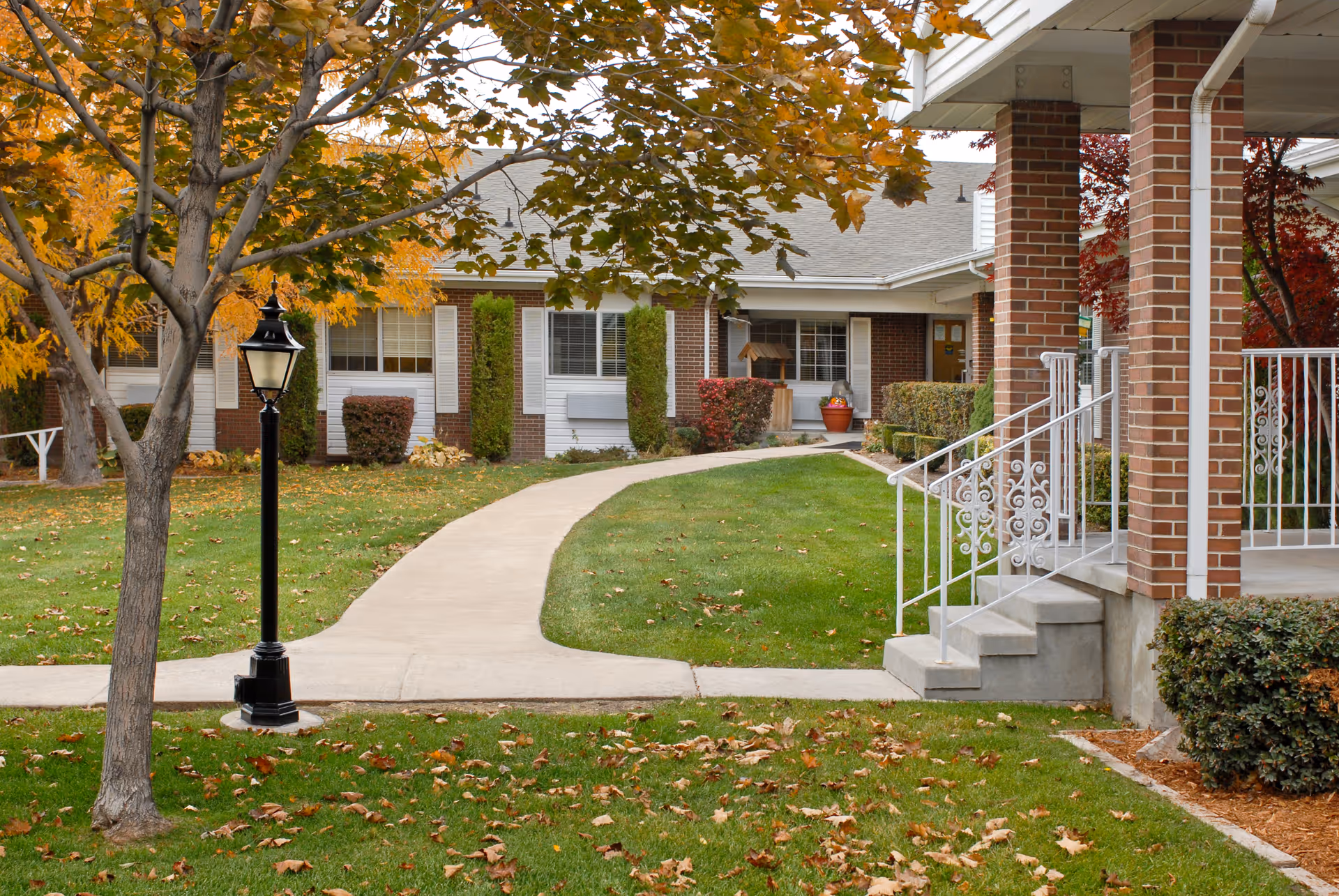 Outdoor view of a senior living facility with a curved concrete walkway, green lawn, autumn trees with orange and yellow leaves, a black lamp post, and a brick building with white window shutters and a porch with white railings and steps.