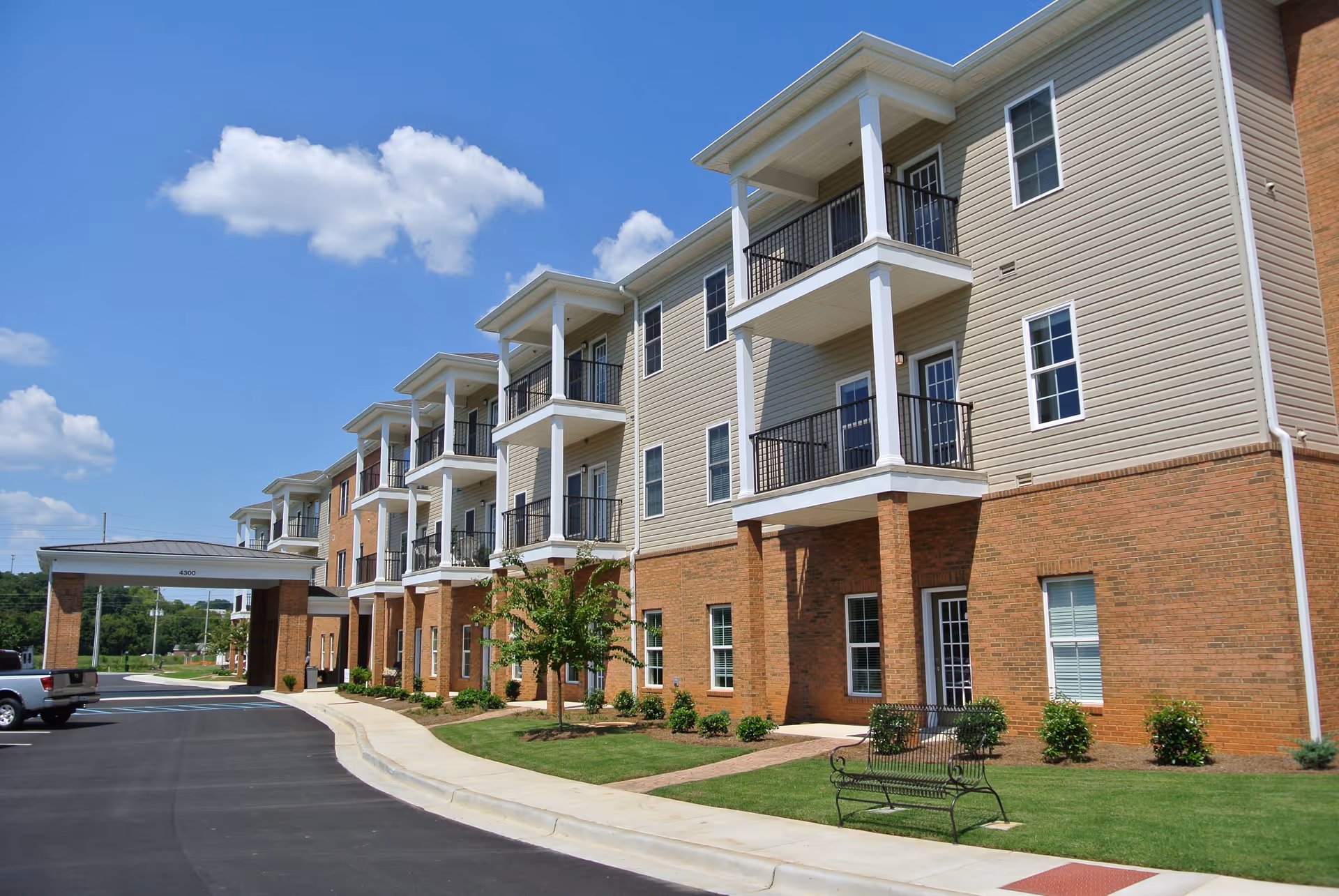 Exterior view of a multi-story residential building with balconies, brick and siding facade, a covered entrance, a parking area, and a bench on a grassy lawn under a blue sky with some clouds.