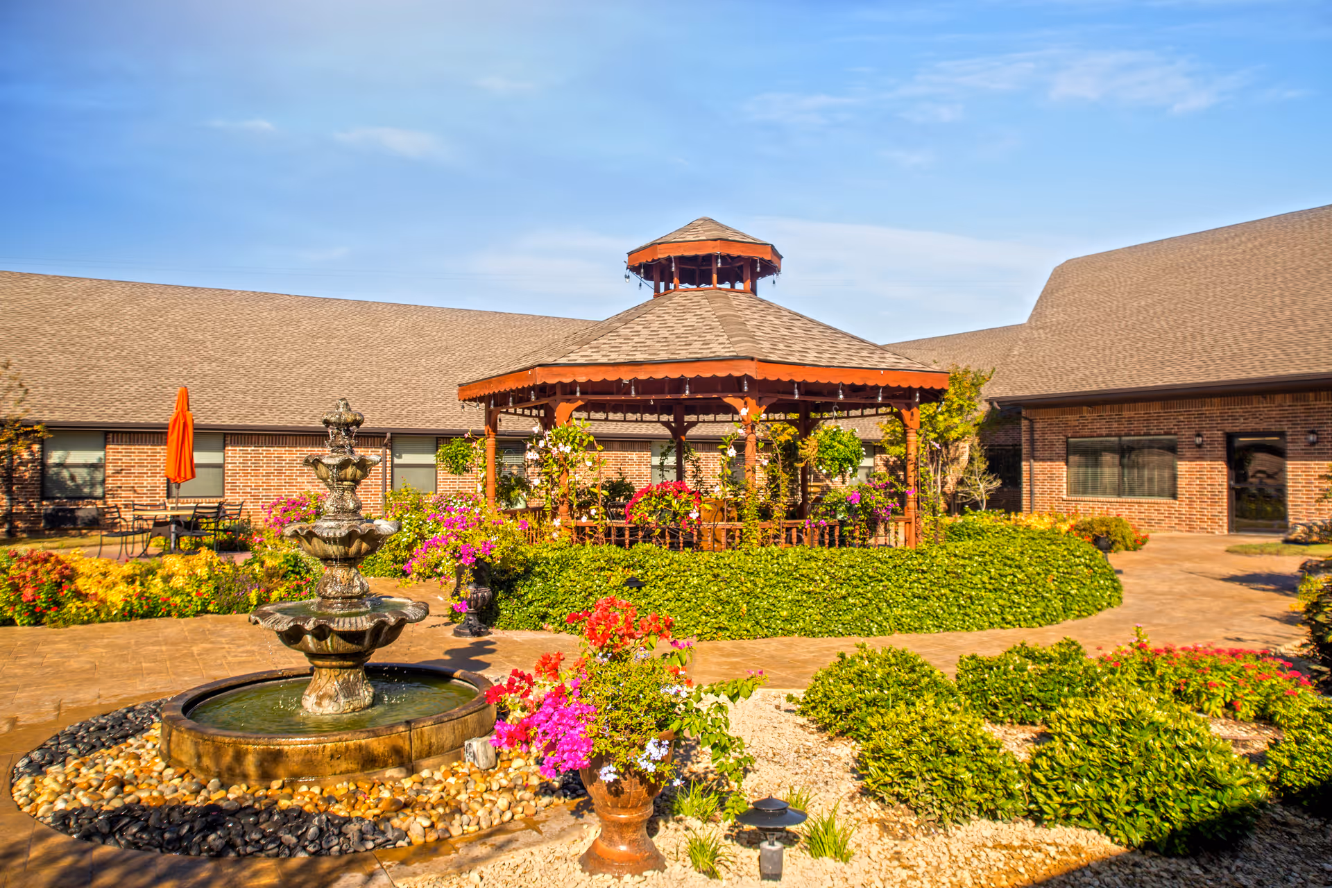 Outdoor courtyard area at Sandy Lake Rehabilitation and Care Center featuring a multi-tiered water fountain surrounded by colorful flowers and greenery, with a wooden gazebo in the background and brick buildings under a clear blue sky.
