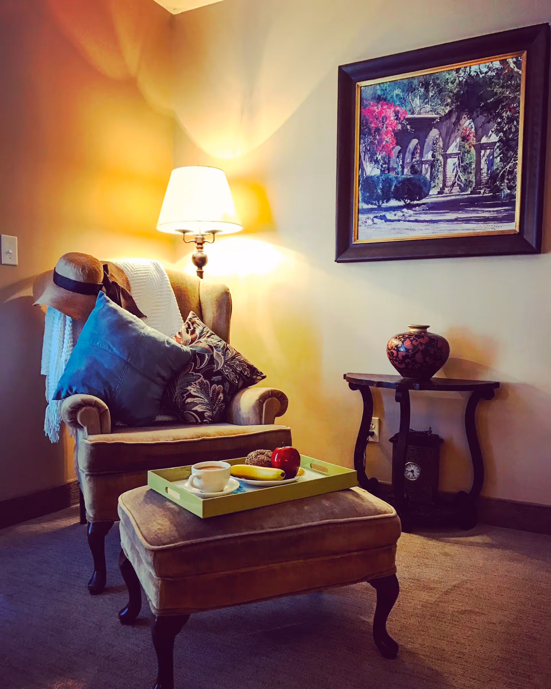 A cozy corner in a living room featuring a cushioned armchair with pillows and a hat hanging on the side, a matching ottoman with a tray holding a cup, a banana, an apple, and a muffin. A lit table lamp is positioned behind the chair, and a framed painting of an outdoor scene hangs on the wall above a small decorative table with a vase and a clock underneath.