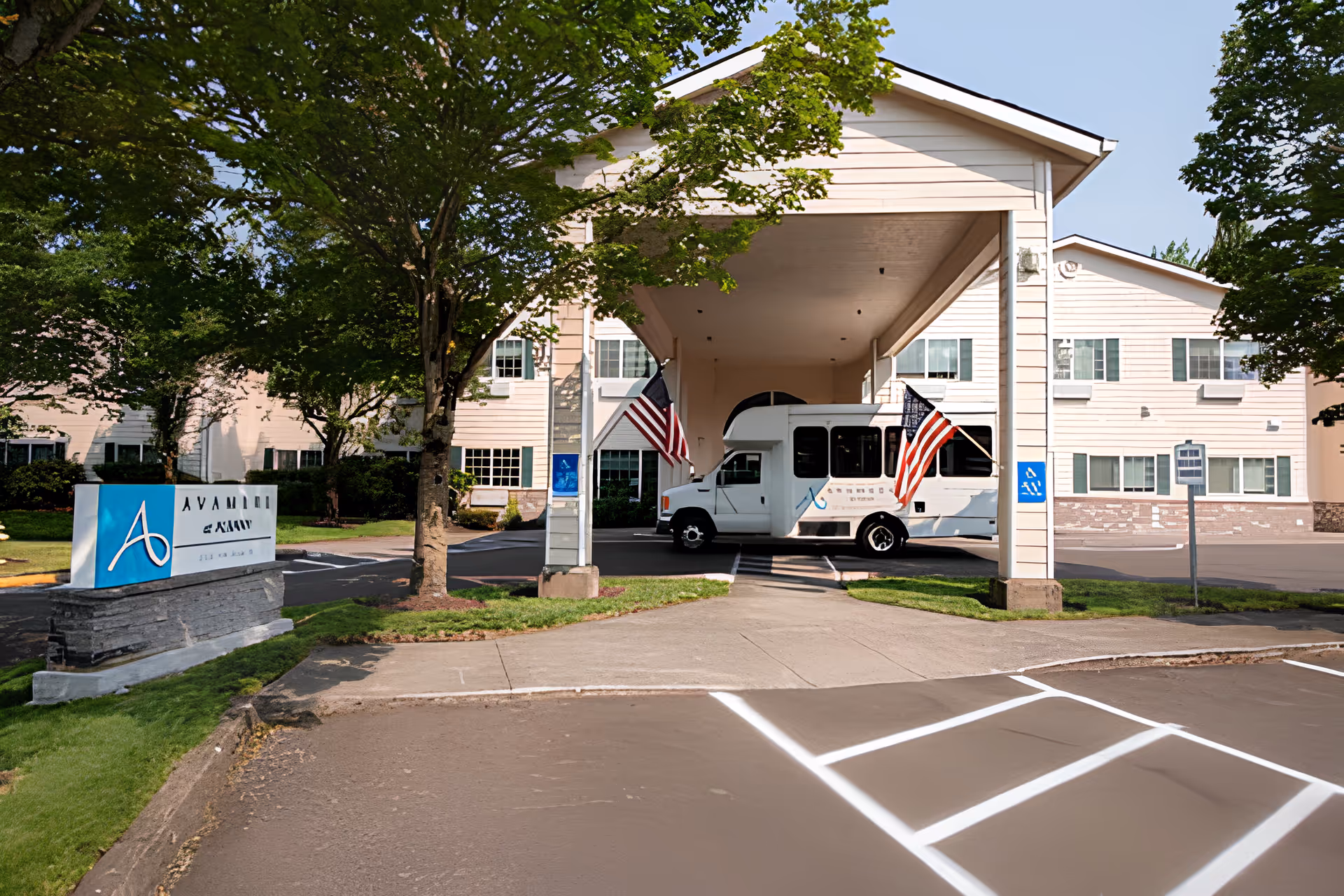 Exterior view of Albany Senior Living by Cogir showing the entrance with a covered drop-off area. A white shuttle bus with American flags is parked under the canopy. The building is light-colored with multiple windows, surrounded by green trees and a well-maintained lawn. A sign with the facility's name is visible near the entrance.