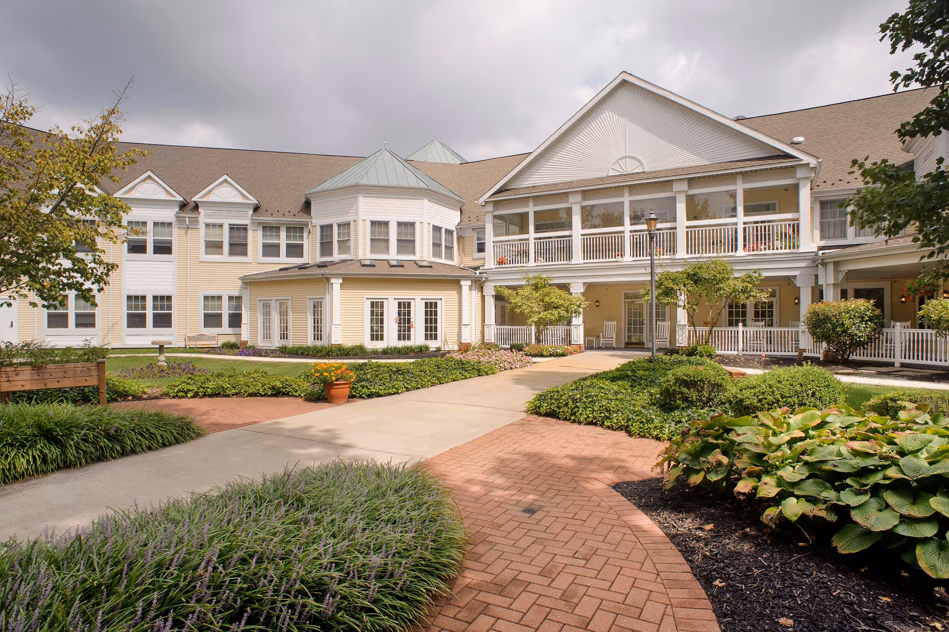 Exterior view of a senior living facility with a well-maintained garden, paved walkways, and a two-story building featuring large windows and a covered porch area with white railings.