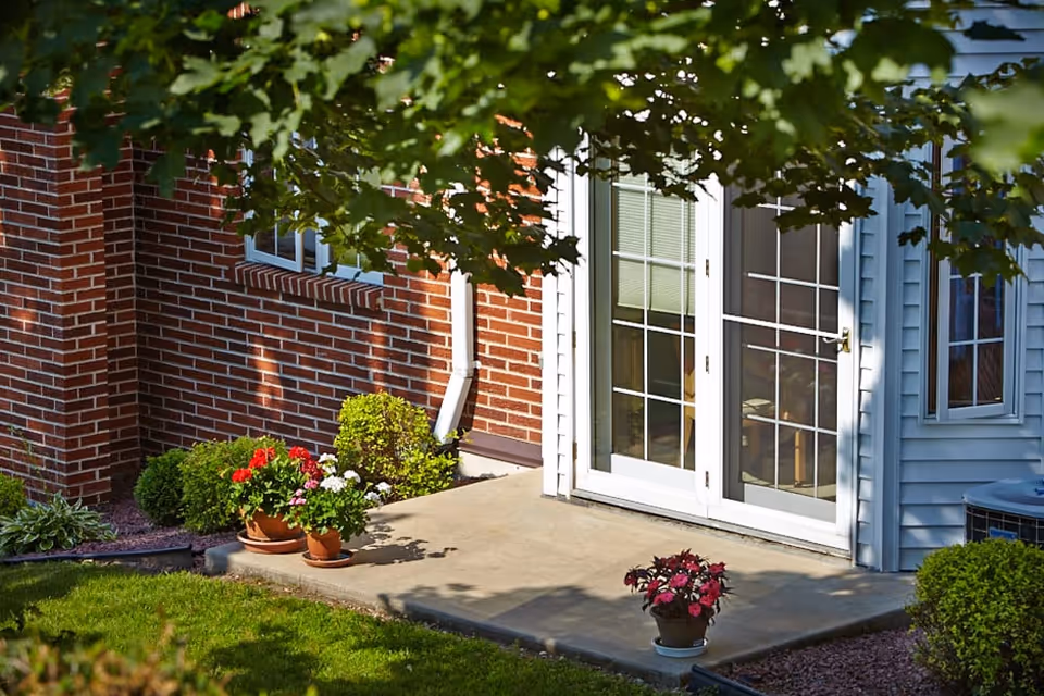 A small concrete patio outside a building with white-framed glass doors and windows. The building has red brick and light blue siding. There are potted plants with colorful flowers on the patio and green bushes around the area. Tree branches with green leaves partially shade the scene.