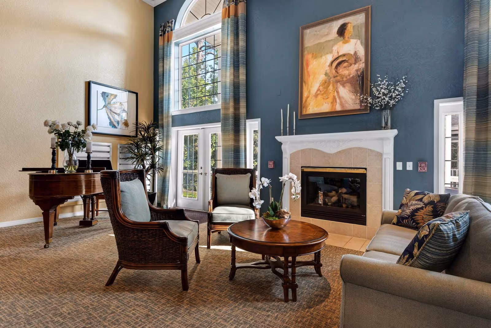 A cozy living room with a beige sofa adorned with patterned cushions, two wooden armchairs with light blue cushions, and a round wooden coffee table with a white orchid centerpiece. The room features a fireplace with a white mantel and a large painting above it. Tall windows with blue and beige striped curtains allow natural light to fill the space. A grand piano and a framed artwork are visible on the left side of the room.