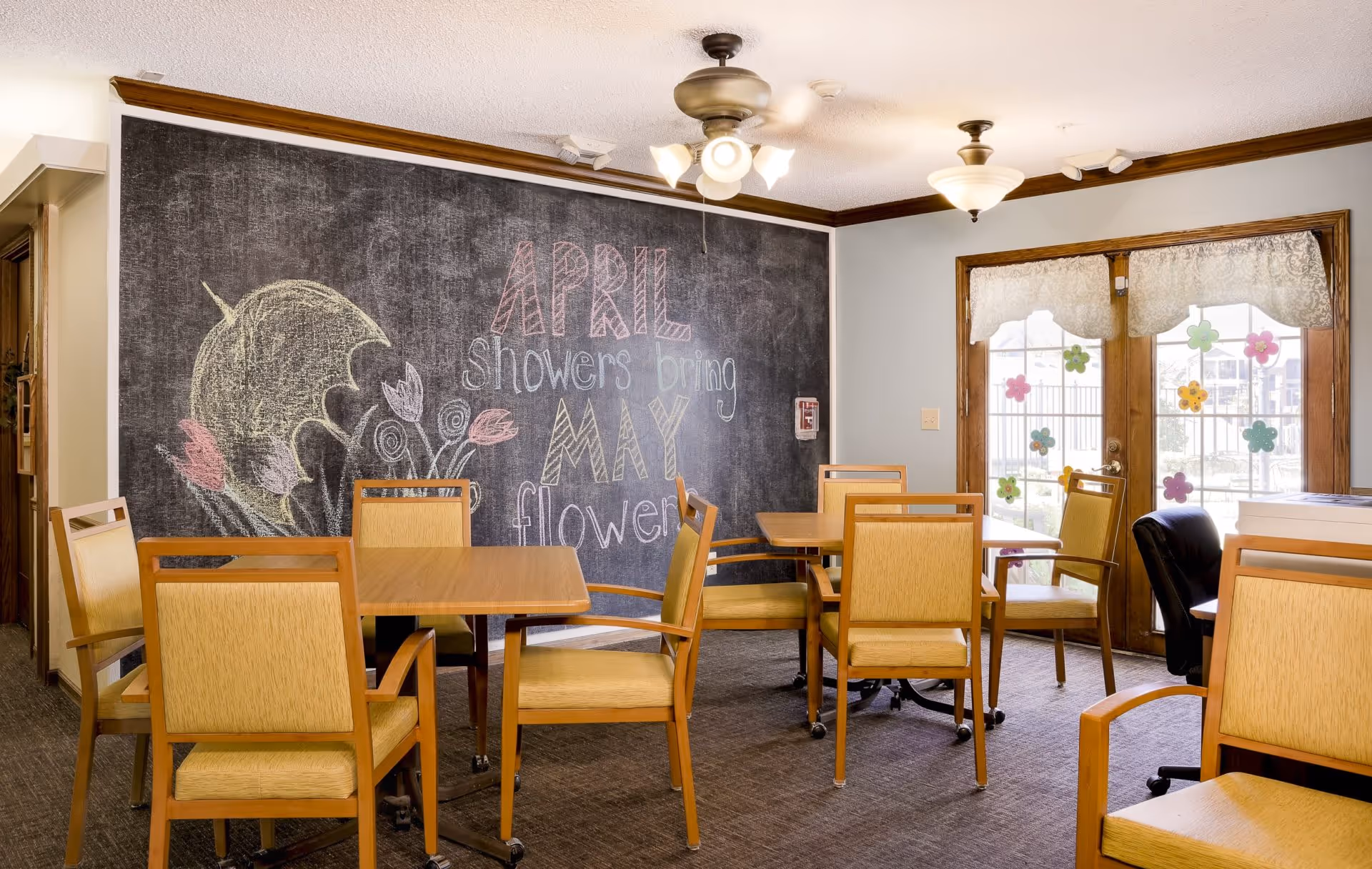 Room with several wooden tables and chairs, a large chalkboard wall reading April showers bring May flowers, and glass doors decorated with paper flowers.