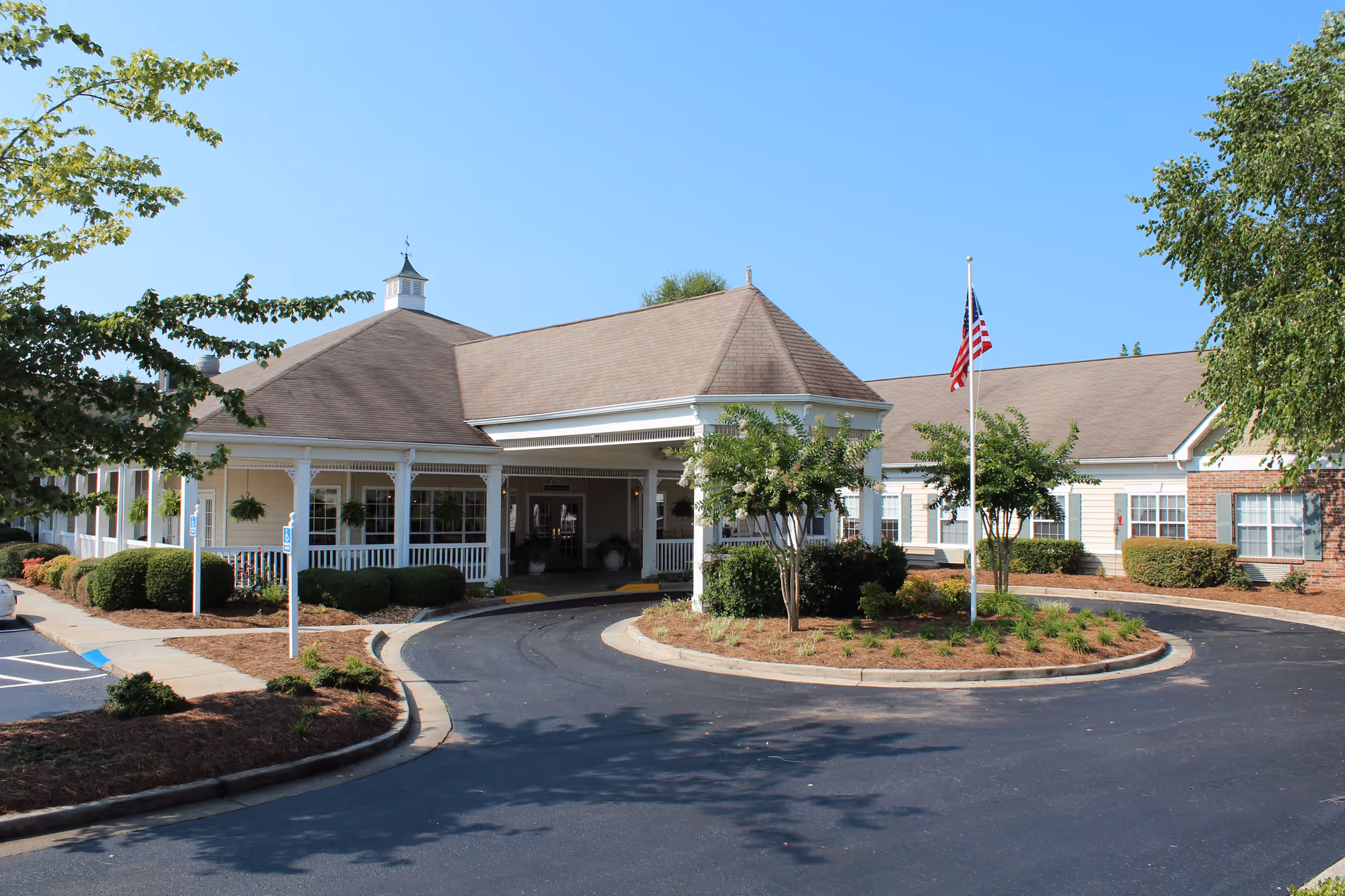 Front exterior of a single-story assisted living facility with a covered porte-cochère, circular driveway, landscaping, and an American flag.