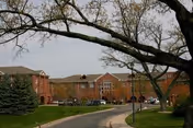 View of the exterior of a brick senior living facility building with a curved driveway, parked cars, and trees with budding leaves in the foreground.
