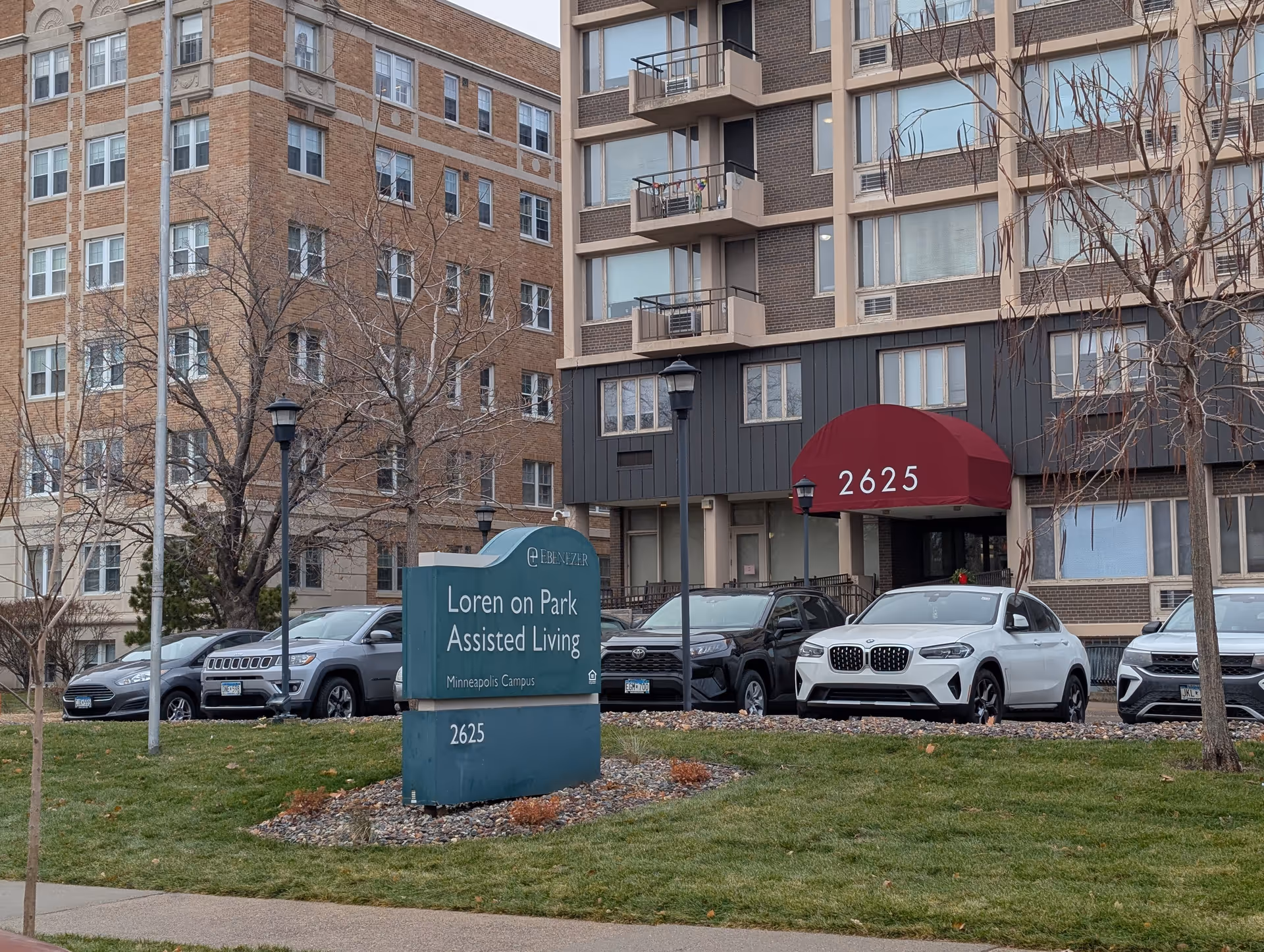 Exterior view of Loren on Park Assisted Living facility with a green sign displaying the name and address 2625. Several parked cars are visible in front of the building, which has multiple windows and balconies. Leafless trees and a grassy area are in the foreground.