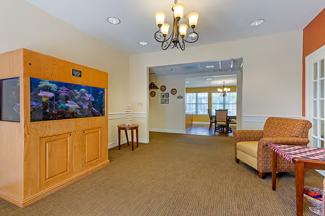 Interior view of a senior living facility hallway with a large built-in aquarium on the left, a small table with a Jenga game, an armchair with a patterned throw on the right, and a dining area visible through an open doorway in the background. The space is well-lit with ceiling lights and a chandelier.