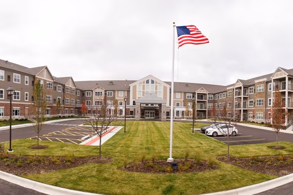 Front exterior of a three-story senior living facility with an American flag on a flagpole in a landscaped courtyard and parking areas.