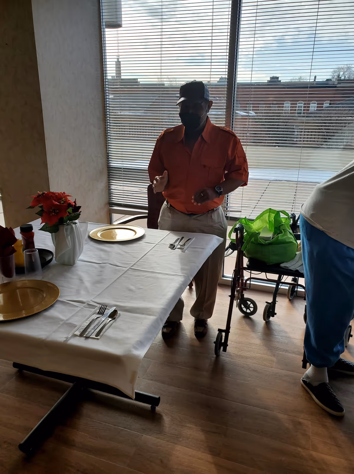 An elderly man wearing a black face mask, orange shirt, and beige pants stands next to a table set with gold plates and silverware in a room with large windows covered by blinds. A walker with a green bag is nearby, and part of another person wearing blue pants and black shoes is visible on the right.