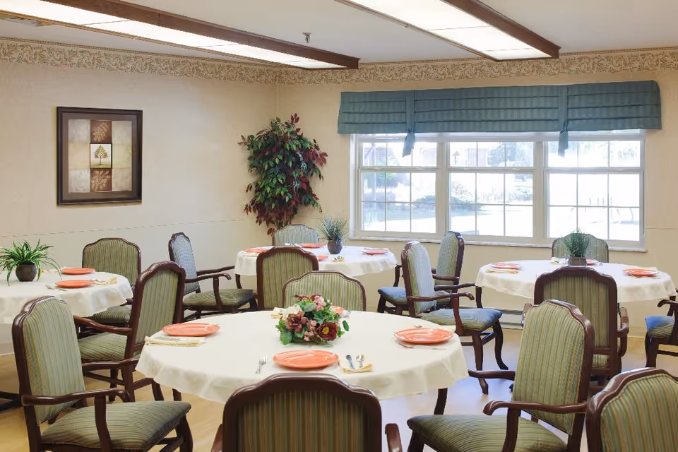 A dining room with round tables covered in white tablecloths, each set with orange plates, napkins, and silverware. The room has green upholstered chairs with wooden frames, a large window with blue valances, a framed artwork on the wall, and a potted plant in the corner.