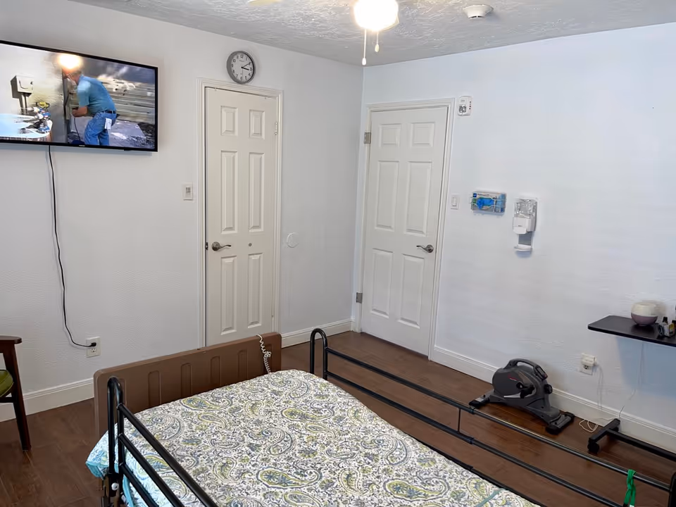 A senior living facility bedroom with a hospital-style bed covered in a patterned bedspread. The room has two closed white doors, a wall-mounted TV showing a person working, a clock above one door, a small exercise pedal machine on the floor, a wall-mounted hand sanitizer dispenser, and a small black shelf with items on it. The floor is wooden and the walls are white.