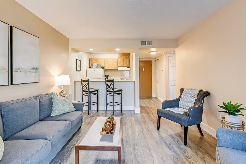 Bright furnished living room with a sofa, armchair, coffee table, and a breakfast bar opening to a small kitchen.