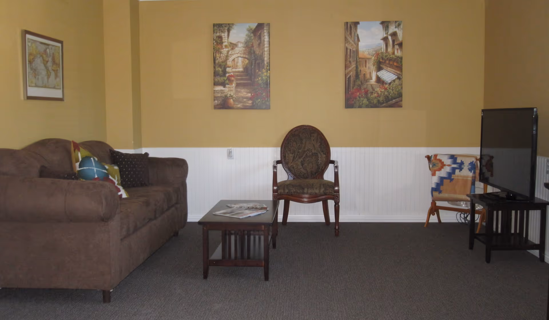 A cozy living room with a brown sofa adorned with colorful pillows on the left, a wooden coffee table with magazines in the center, an ornate wooden chair with patterned upholstery against a beige and white wall, two framed paintings of outdoor scenes hanging above, and a flat-screen TV on a black stand to the right with a small chair covered by a colorful blanket behind it.