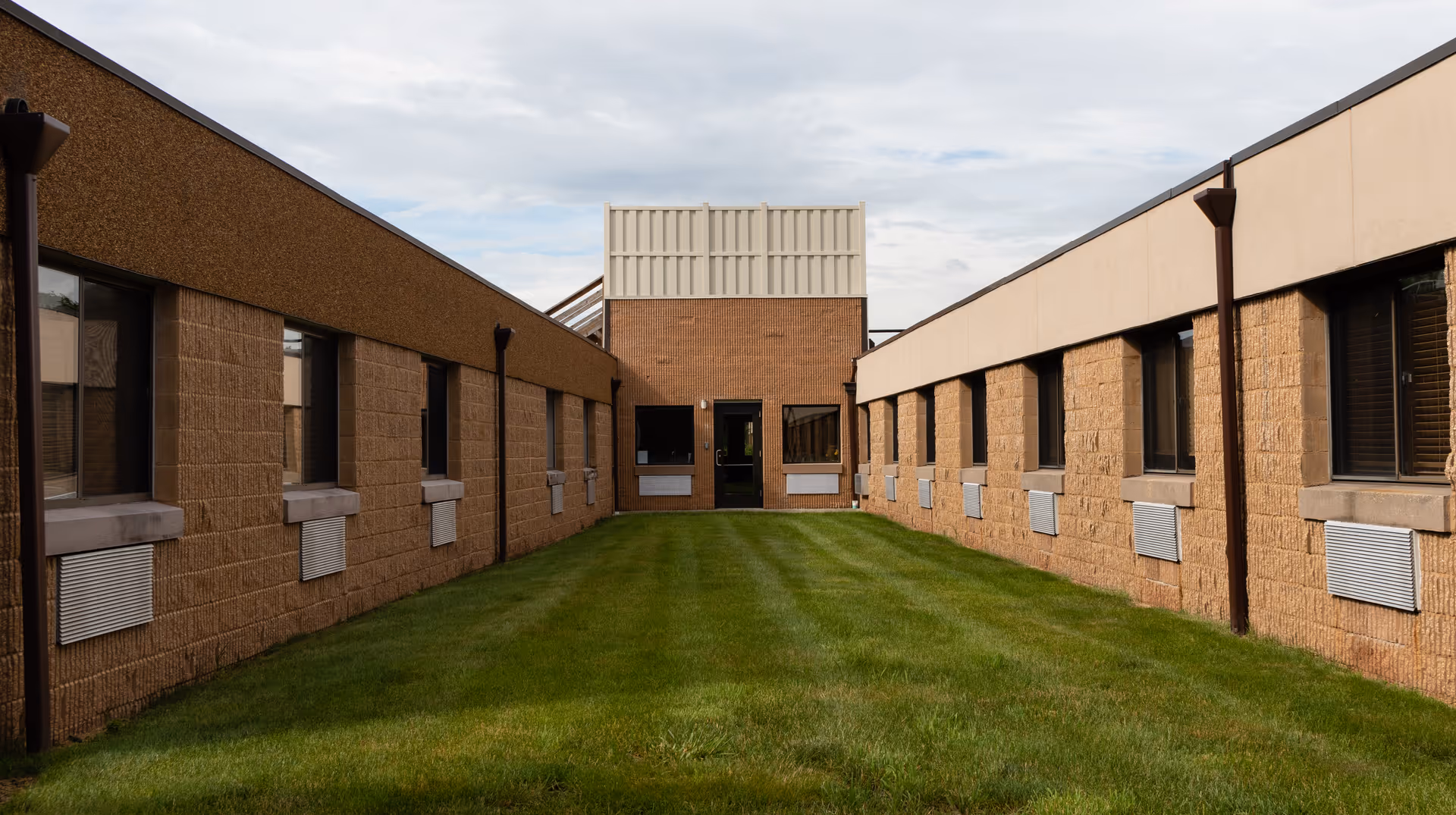 A grassy courtyard area between two single-story brick buildings with multiple windows, under a cloudy sky.