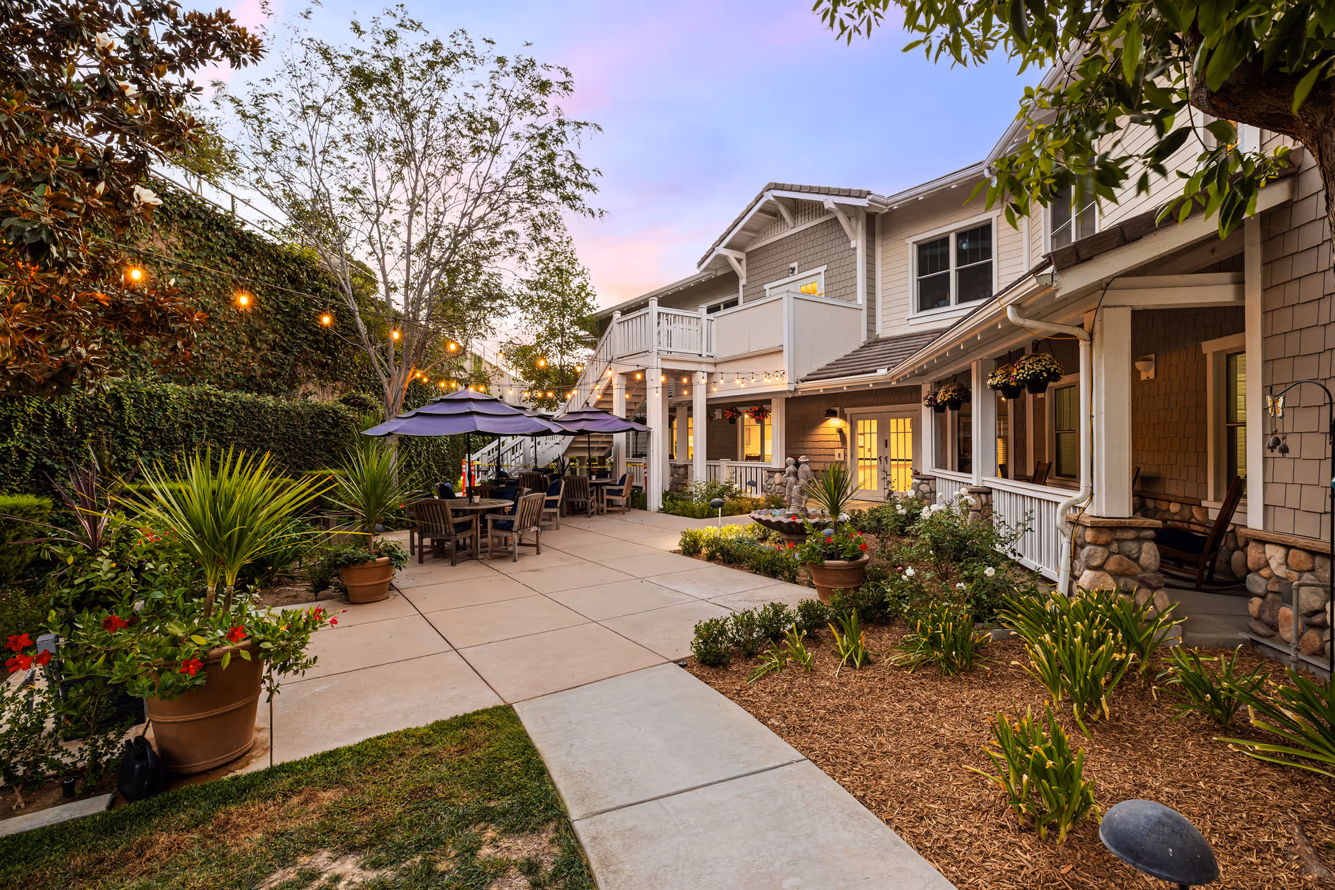 Outdoor patio area at Ivy Park at Wood Ranch with tables and chairs under umbrellas, string lights hanging above, surrounded by plants and greenery, adjacent to a two-story building with a porch and balcony during sunset.