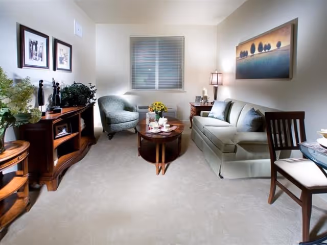 A cozy living room with a green upholstered sofa, a matching armchair, a wooden coffee table with a tea set and flowers, a wooden side table with a lamp, framed pictures on the wall, and a window with blinds.