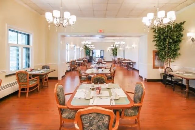 A spacious dining room with multiple tables and chairs arranged neatly. The tables are set with placemats, cups, and utensils. The room features wooden flooring, large windows with blinds, chandeliers hanging from the ceiling, and potted plants along the walls.