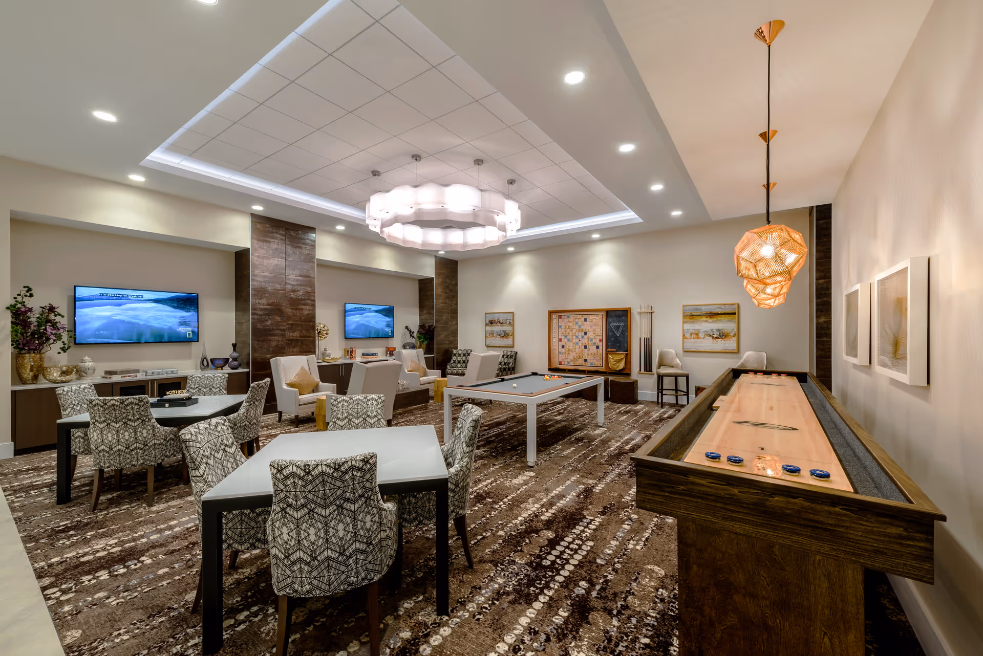 A spacious and well-lit recreational room with patterned carpet flooring. The room features a shuffleboard table on the right, a pool table in the center, and two tables with patterned chairs on the left. At the back, there are two wall-mounted televisions above cabinets, several armchairs, and decorative items. The ceiling has recessed lighting and a large modern chandelier in the center.