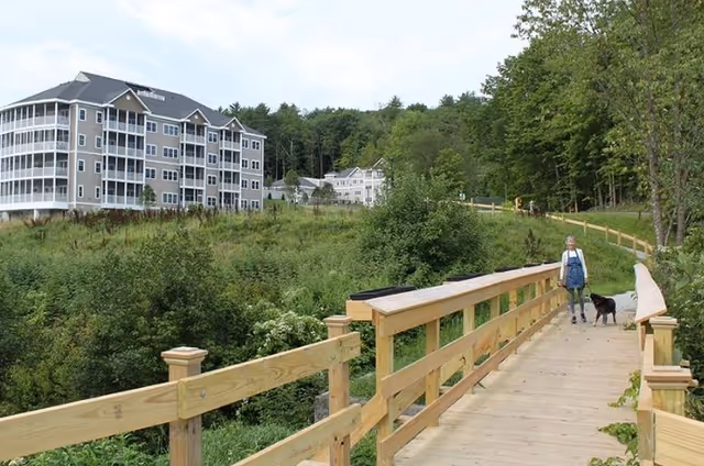 A senior woman walking a black dog on a wooden bridge surrounded by greenery with multi-story residential buildings in the background under a partly cloudy sky.