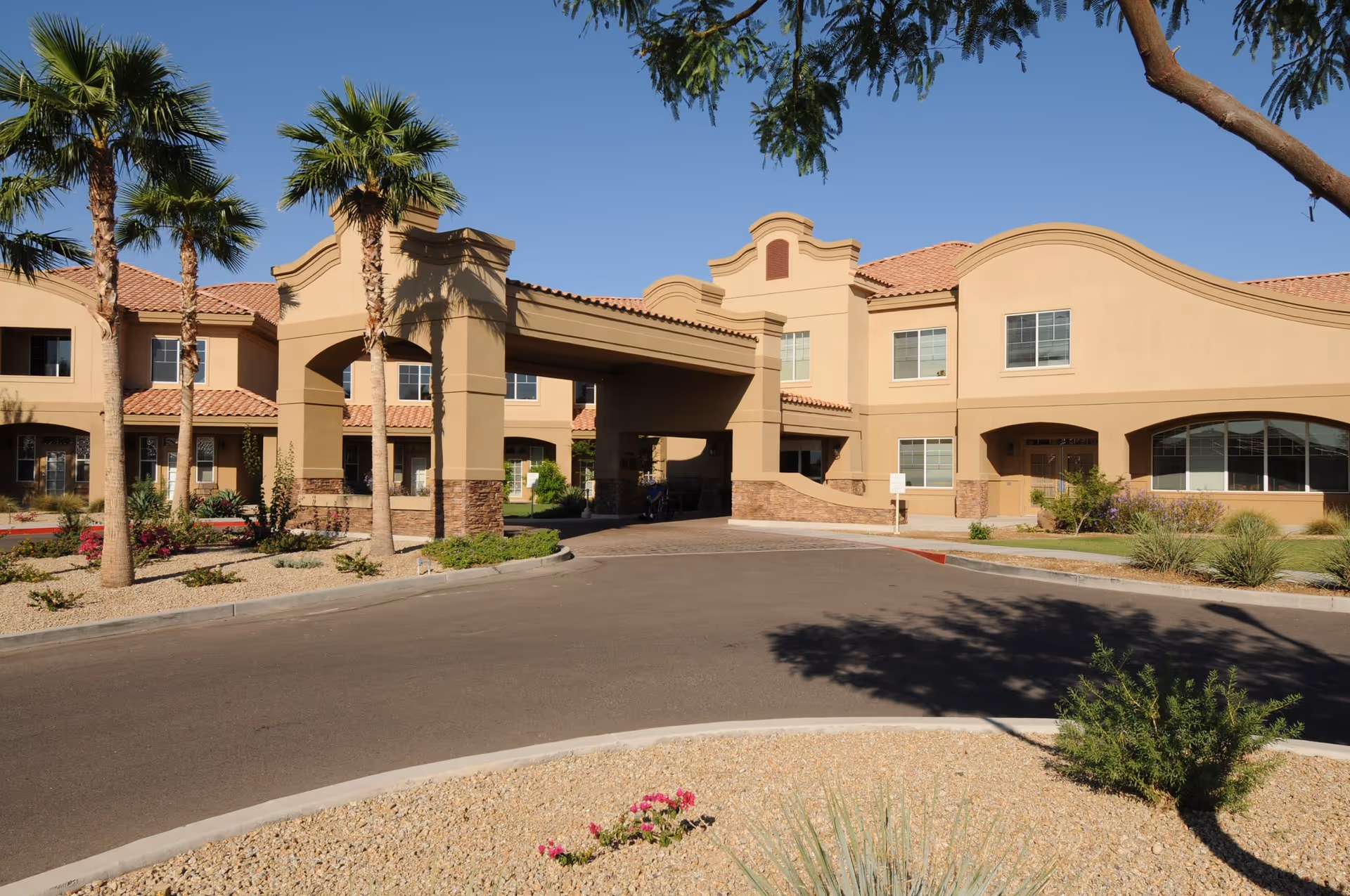 Exterior view of La Loma Village Health & Rehabilitation Center showing a beige two-story building with a covered entrance, palm trees, and desert landscaping under a clear blue sky.
