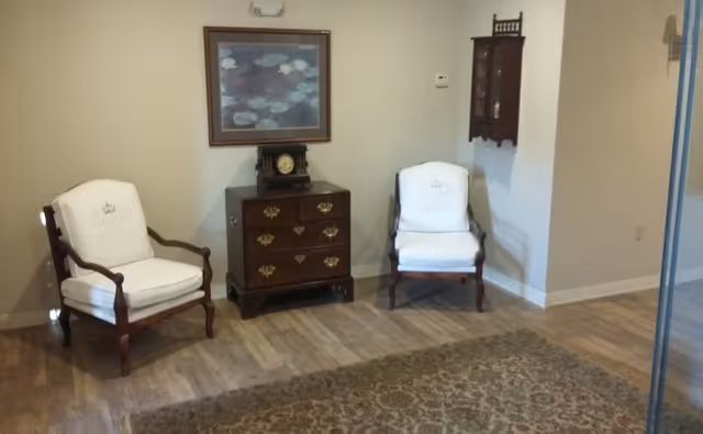 Two upholstered armchairs flank a small wooden chest with a clock under framed wall art in a sitting area with a patterned rug.