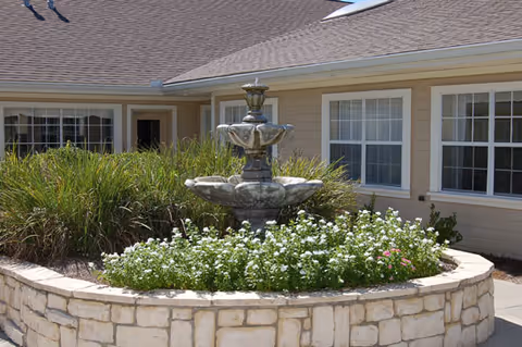Outdoor stone fountain surrounded by white flowers and greenery in front of a beige building with multiple windows and a shingled roof.