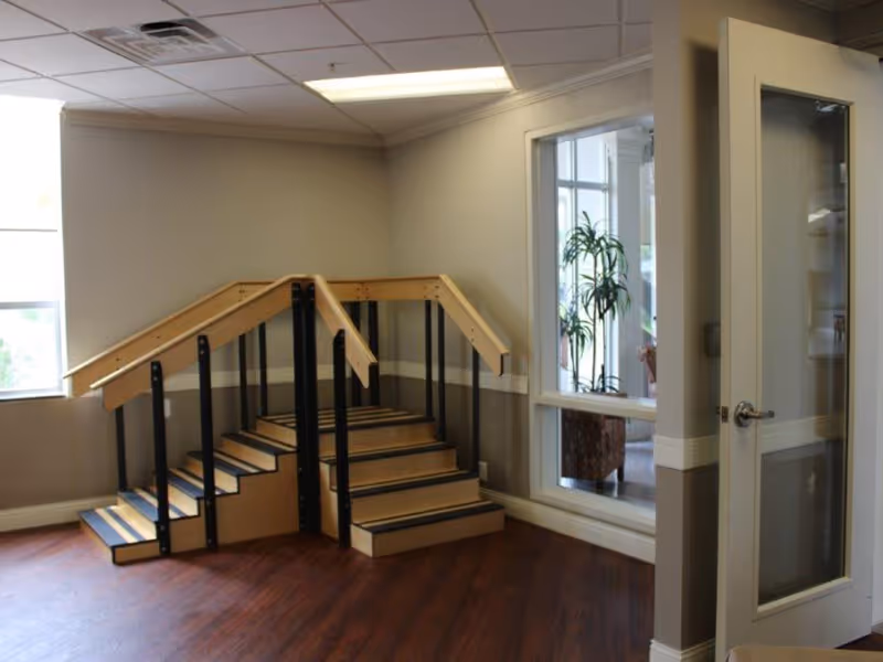 A small indoor practice staircase with handrails in a rehab facility room with wood floors and a door opening to an adjacent space.