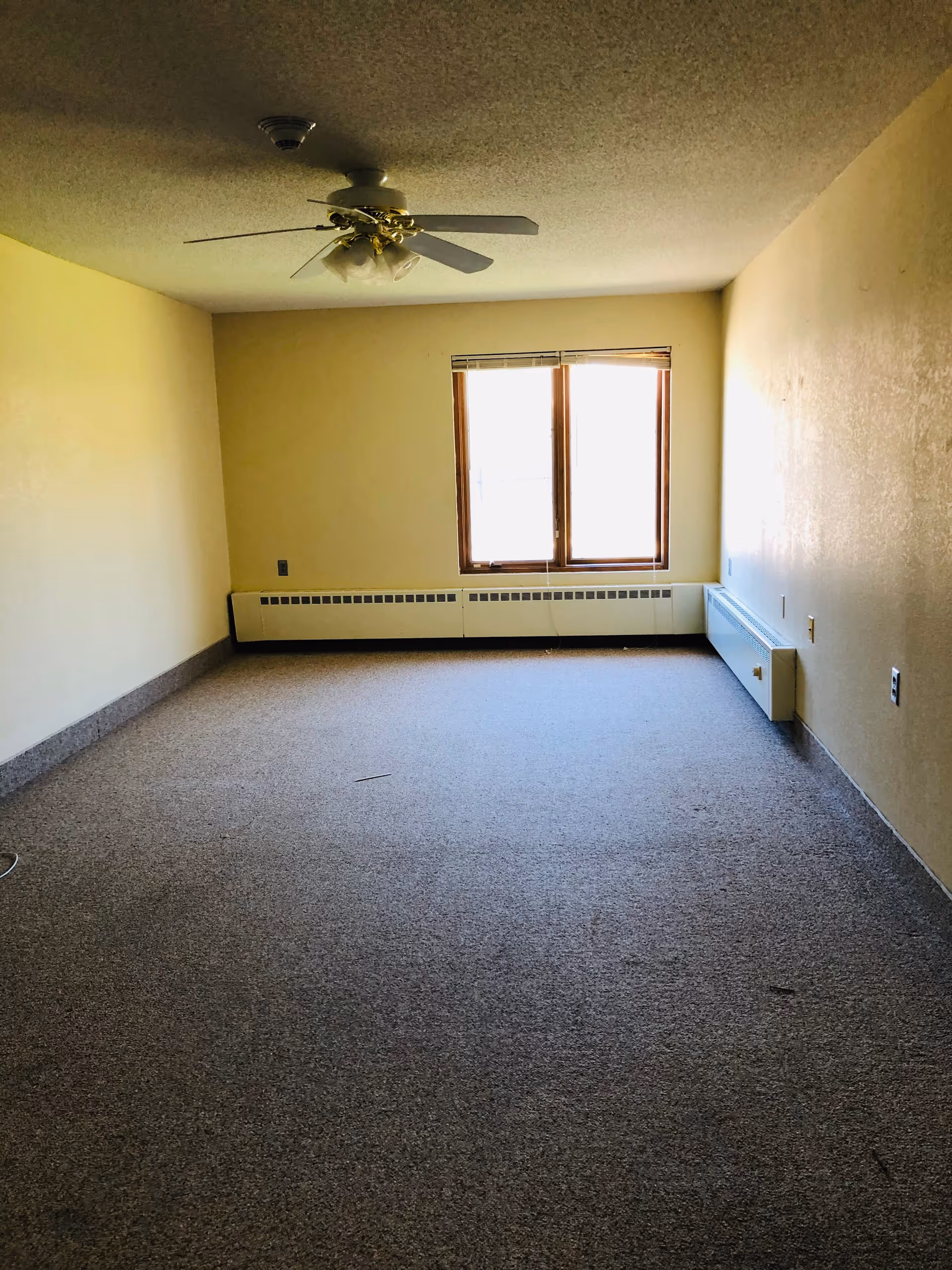 Empty room with beige walls and carpeted floor, a ceiling fan with lights, and a window with partially open blinds letting in natural light.