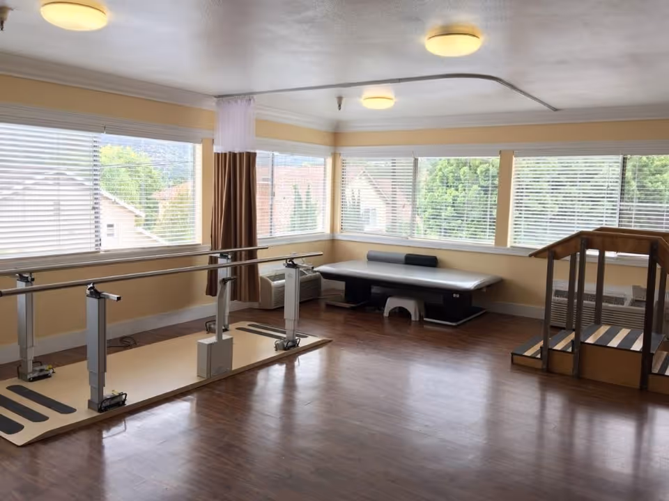 Bright rehabilitation room with parallel bars, an exercise bench and practice stairs beside large windows.
