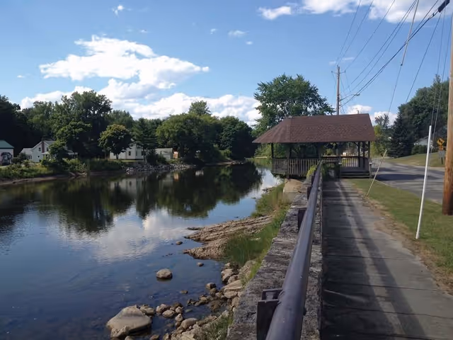 A peaceful riverside scene with a calm river reflecting the blue sky and clouds. On the right side, there is a paved walkway with a metal railing and a wooden gazebo with a shingled roof. Trees and houses are visible in the background along the riverbank.