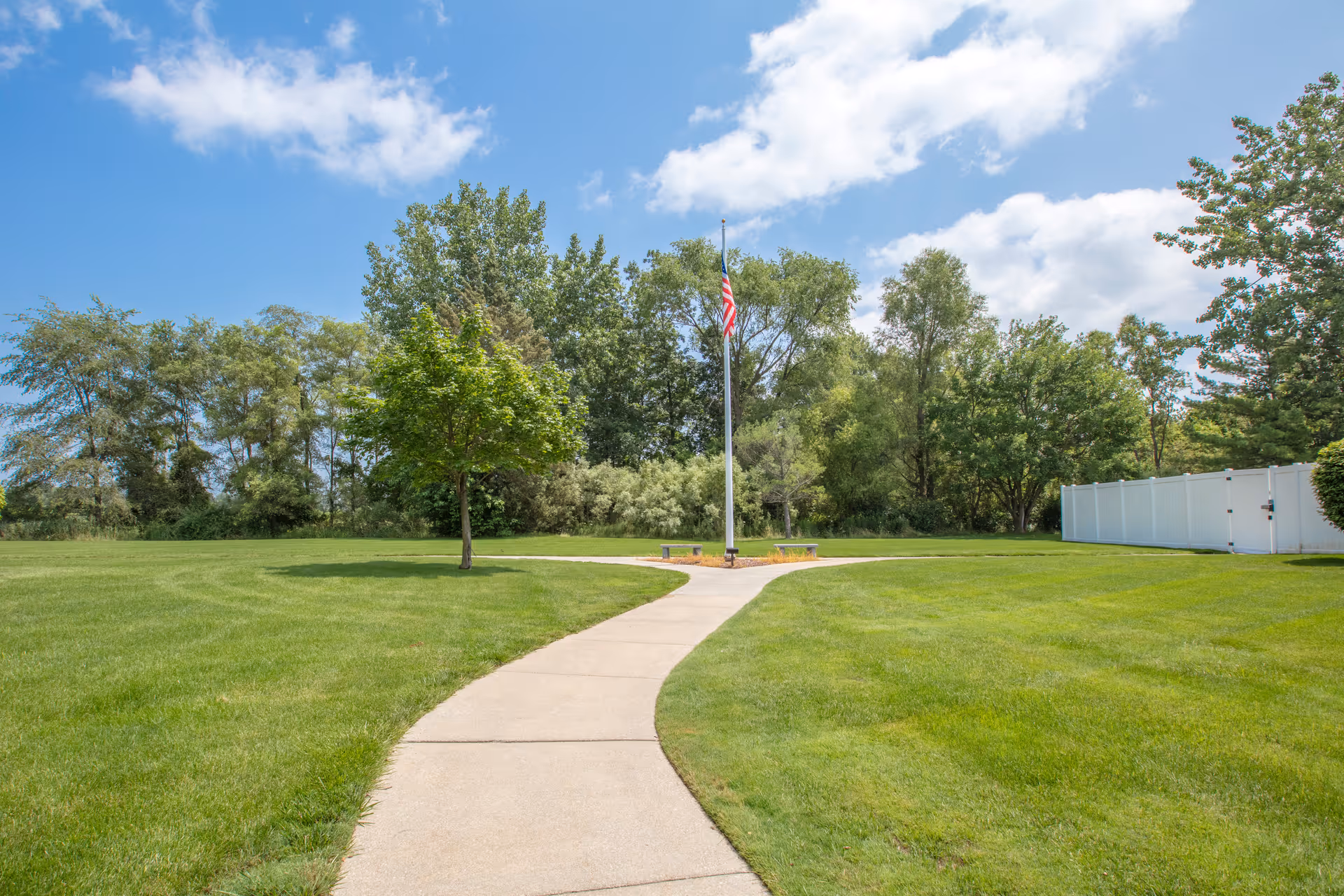 Concrete walkway through a green lawn leading to an American flagpole with trees and a blue sky in the background.