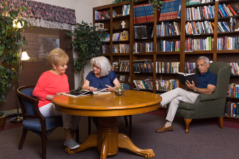 Three people reading and looking at books around a round wooden table in a cozy library-like common room with bookshelves.