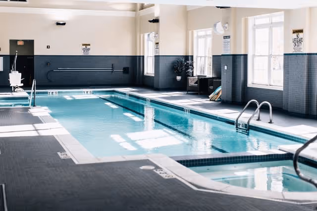Indoor swimming pool area with clear water, large windows letting in natural light, poolside handrails, and safety signs on the walls.
