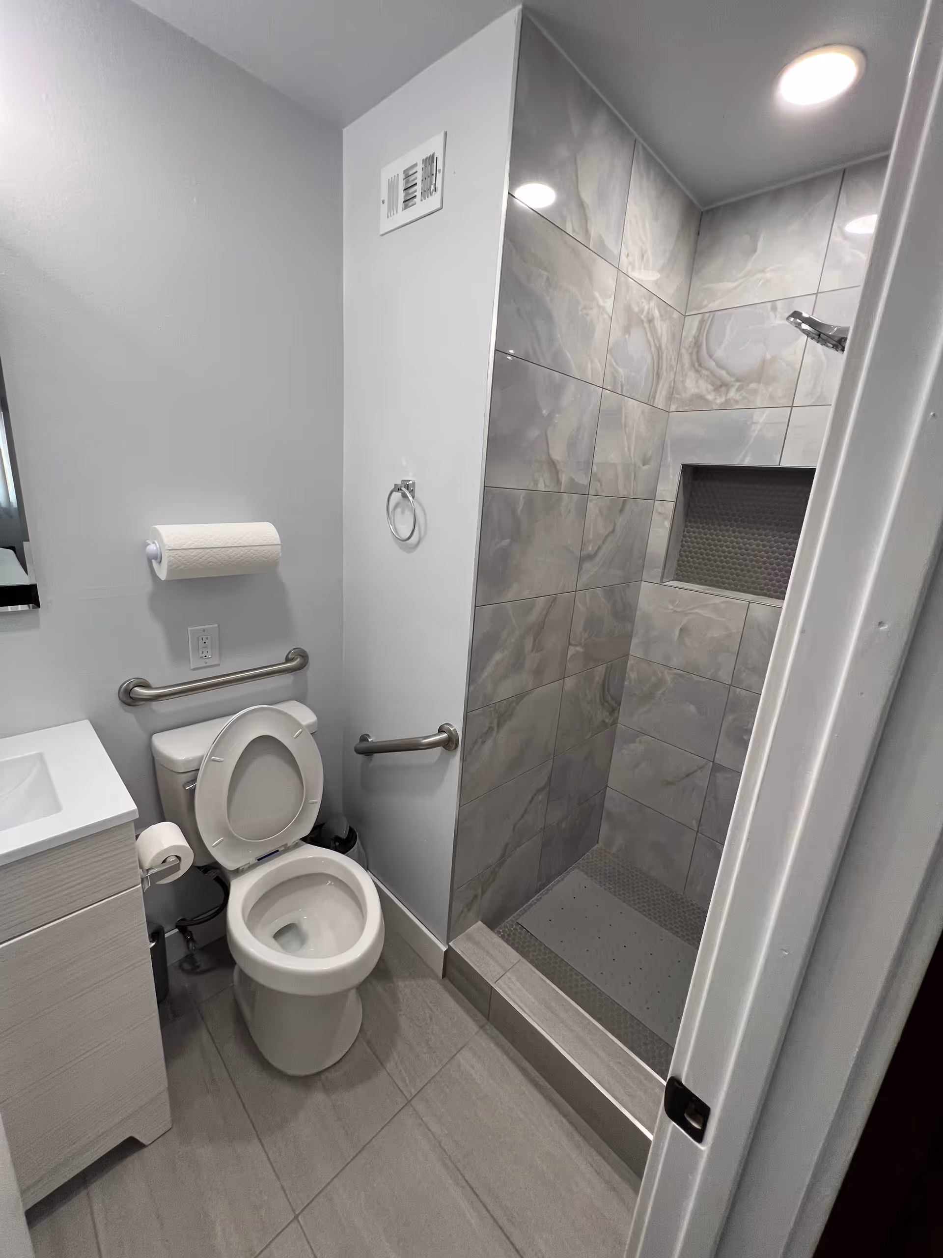 A small bathroom with a white toilet that has the seat up, a white vanity with a sink, and a walk-in shower with gray marbled tiles and a built-in niche. The bathroom has gray floor tiles and white walls, with grab bars installed near the toilet for accessibility.