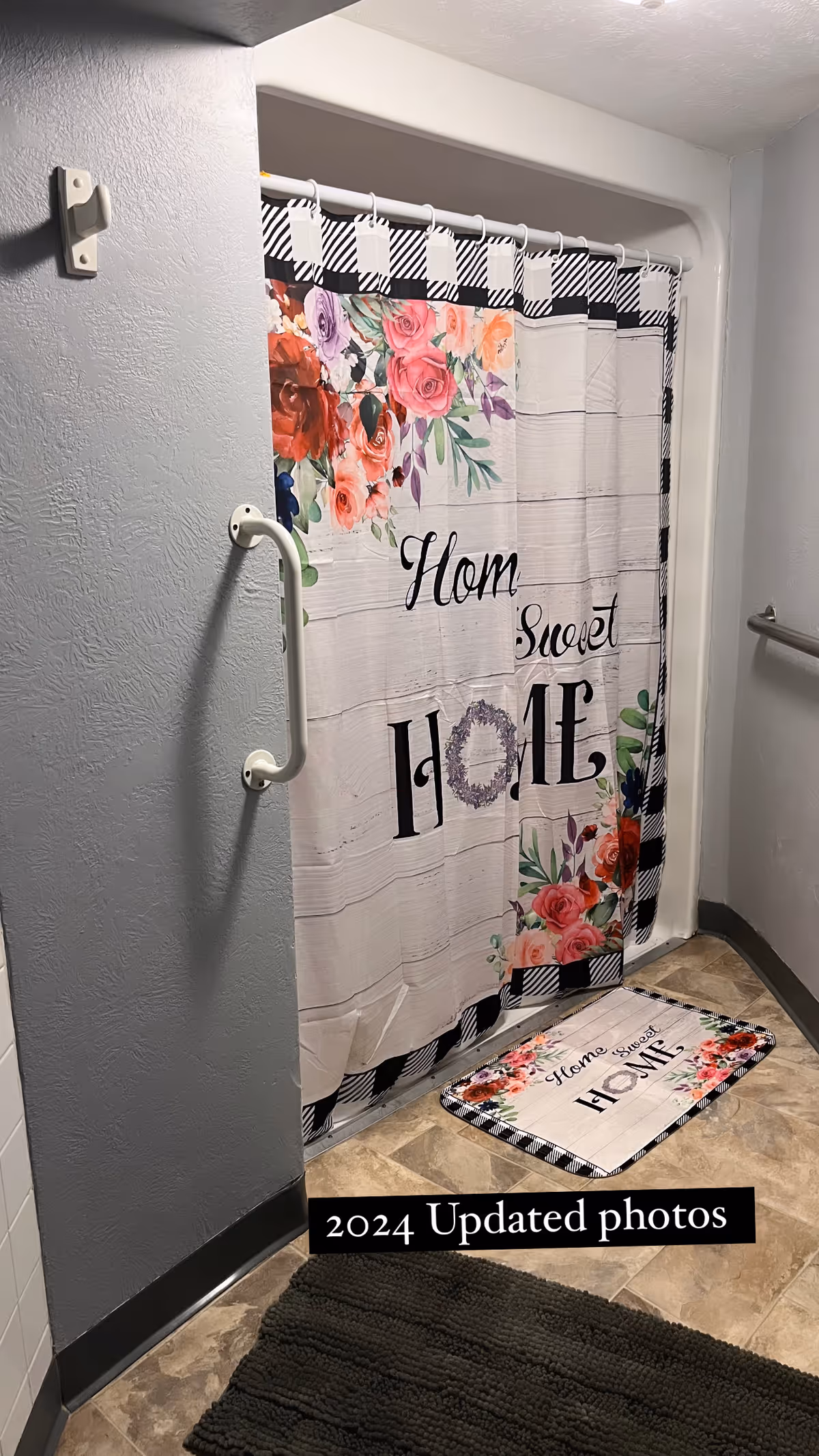 A bathroom shower area with a floral and checkered shower curtain that reads 'Home Sweet Home'. There is a matching floor mat in front of the shower with the same text and floral design. The walls are light gray, and there is a white grab bar mounted on the wall next to the shower. The floor has a beige tile pattern, and a dark gray bath mat is partially visible in the foreground.