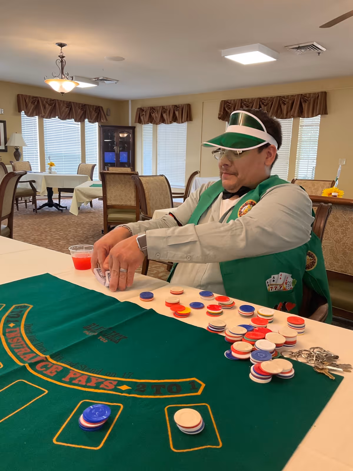 A man wearing a green visor and green vest sits at a table with a green felt blackjack layout, handling playing cards and surrounded by poker chips. The room has beige walls, carpeted floor, several tables and chairs, and windows with brown valances.