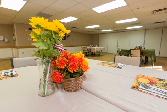 A dining room with tables covered in light-colored tablecloths, decorated with vases of bright yellow sunflowers and orange flowers. The tables have place settings with utensils and colorful placemats. The room has a drop ceiling with fluorescent lights and beige walls with a chair rail.