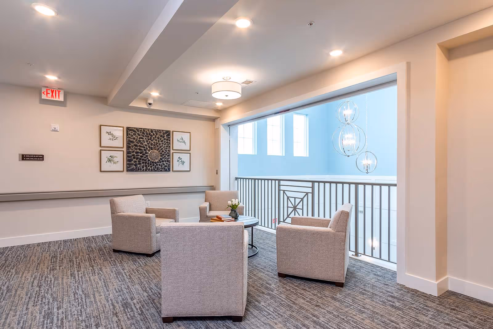 A cozy seating area with four beige armchairs arranged around a small round table with a vase of white flowers and some books. The space has carpeted flooring, light-colored walls, framed botanical artwork on the wall, and a railing overlooking a bright area with large windows and modern hanging light fixtures.