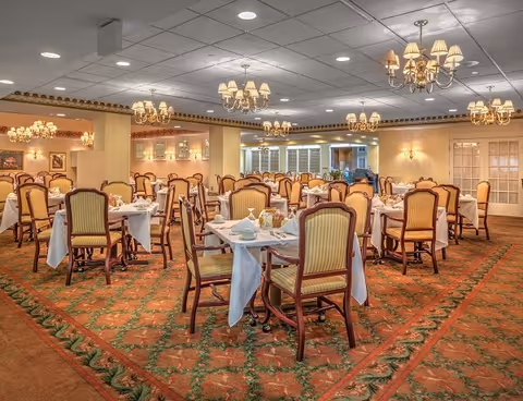 A spacious dining room with multiple tables covered in white tablecloths, set with plates, cups, and silverware. The room features elegant chandeliers hanging from the ceiling, upholstered wooden chairs with striped cushions, and a patterned carpet with floral designs. The walls are painted in warm tones with decorative molding and glass-paneled doors in the background.