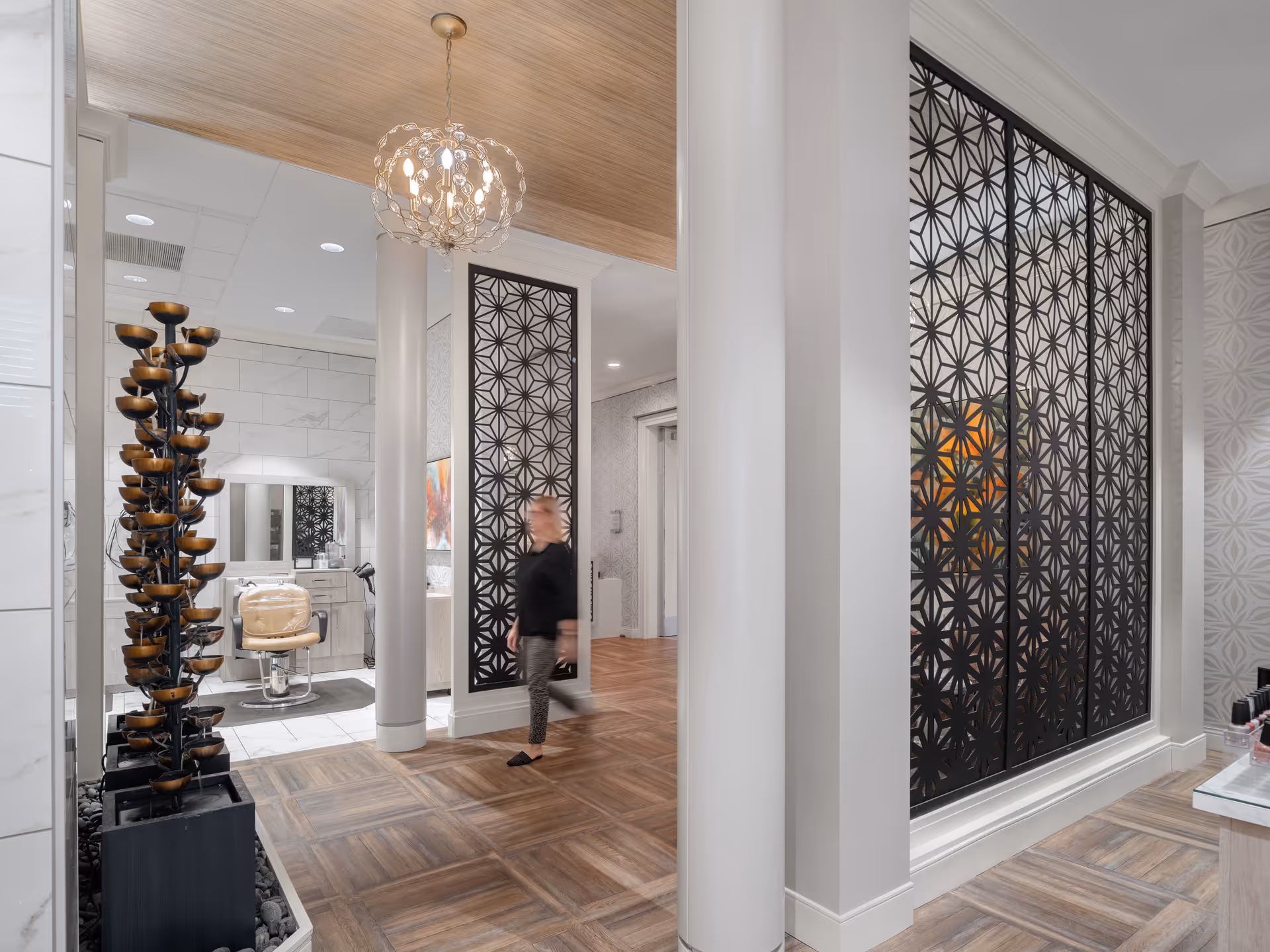 Modern senior living interior hallway featuring decorative black patterned screens, white columns, wood-look flooring, a sculptural fountain, a salon chair, and a blurred person walking.