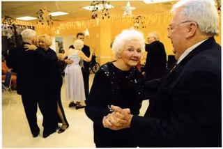 Several elderly couples dressed formally are dancing in a decorated room with hanging star and string lights, creating a festive atmosphere.
