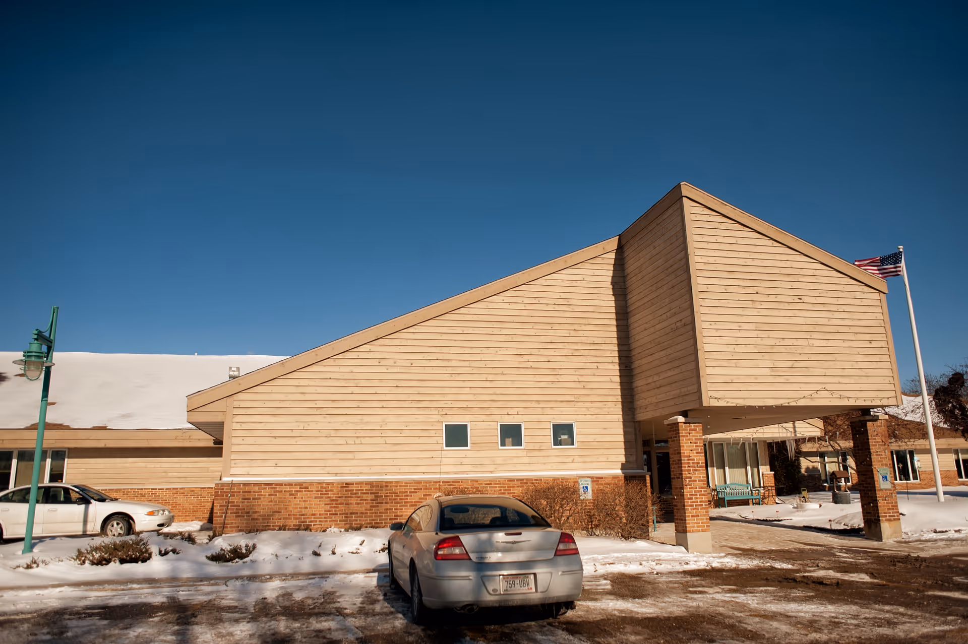 Exterior view of a senior living facility building with a sloped roof and a covered entrance supported by brick pillars. There are two cars parked in front, snow on the ground, a green street lamp on the left, and an American flag on a flagpole to the right under a clear blue sky.