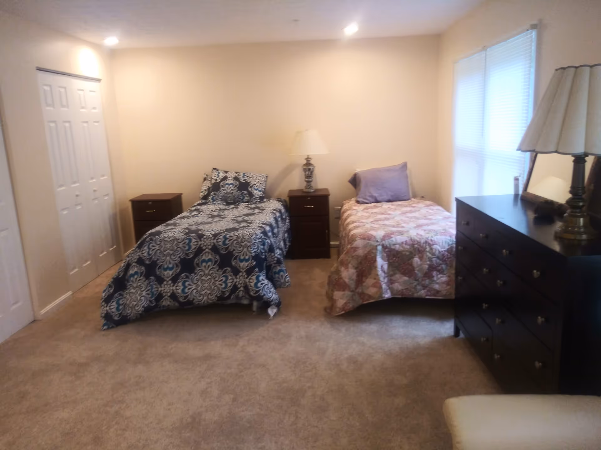 A bedroom with two single beds, each with different patterned bedspreads and pillows. Between the beds is a small nightstand with a lamp. There is a large dresser with a mirror and another lamp on the right side of the room. The room has beige walls, carpeted floor, and a window with blinds.